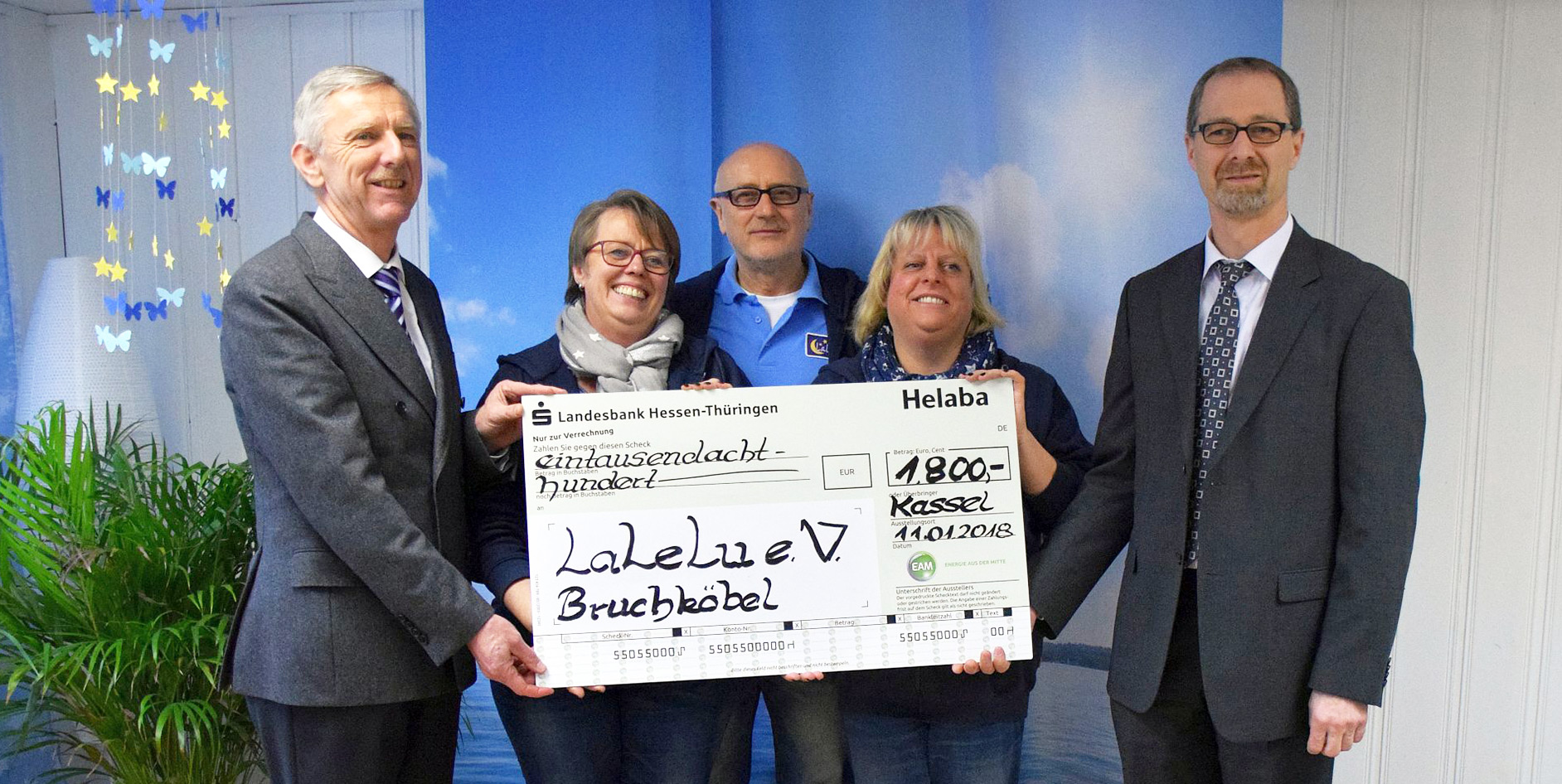 Gruppe von fünf Personen hält einen Scheck über 1.800 Euro für den Verein LaLeLu e.V. vor einer blauen Wand.