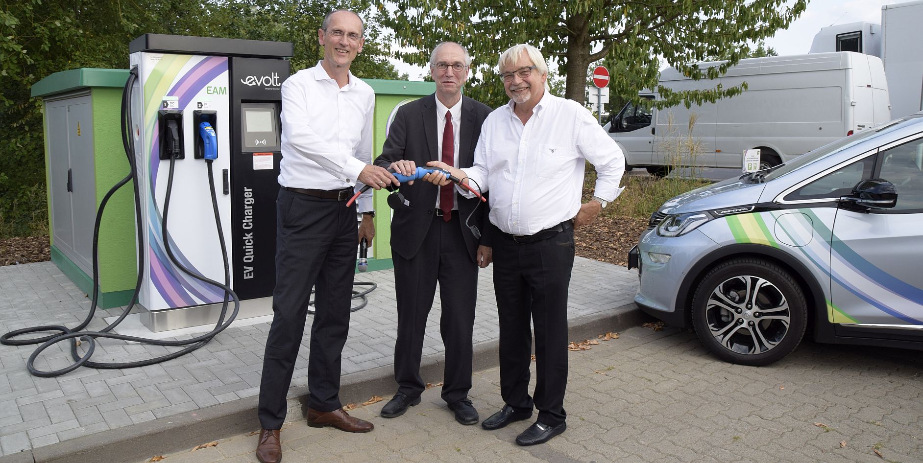 Drei Männer an einer Ladestation für Elektrofahrzeuge halten ein Kabel und lächeln in die Kamera.