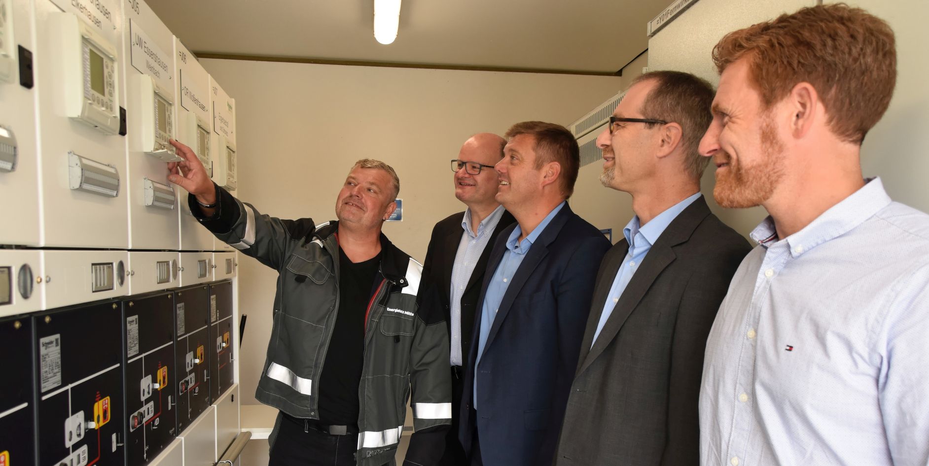 Fünf Männer stehen vor elektrischen Messtechnik-Geräten und diskutieren, während einer auf ein Gerät zeigt.