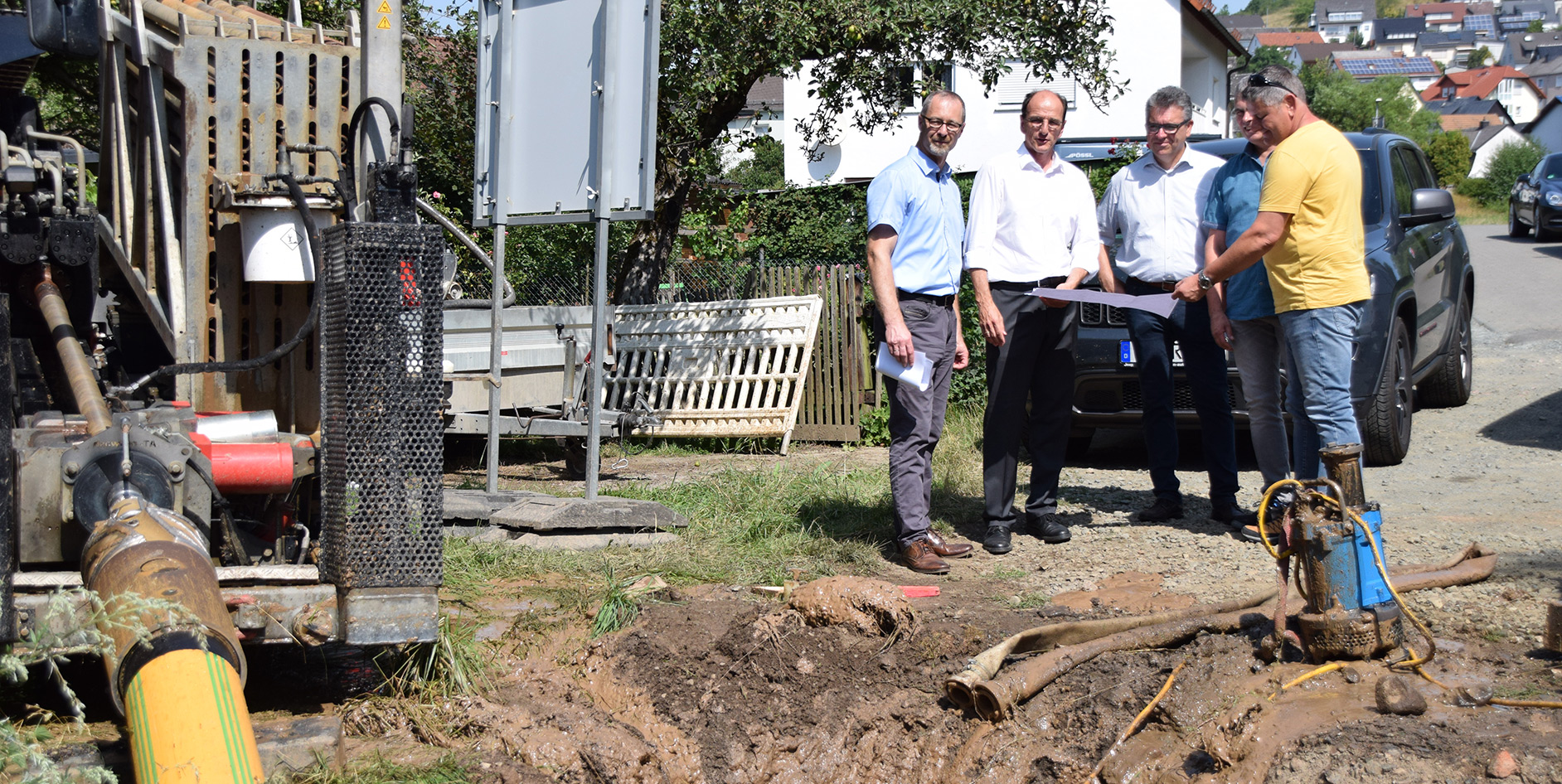 Vier Personen besprechen eine Baustelle neben einem Bagger in einem Wohngebiet, umgeben von Erde und Rohren.