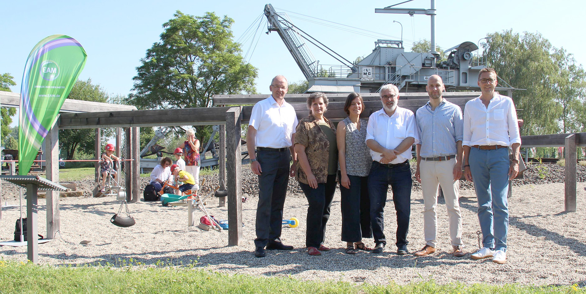 Gruppenszene auf einem Spielplatz mit sechs Personen, einem grünen Banner und spielenden Kindern im Hintergrund.