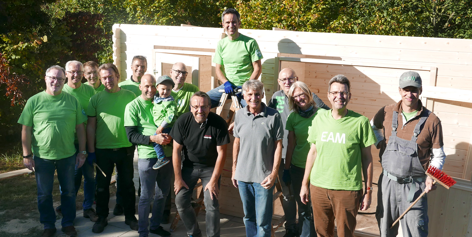 Gruppe von Menschen in grünen T-Shirts, die vor einem Holzbau stehen, lächeln und posieren zusammen im Freien.