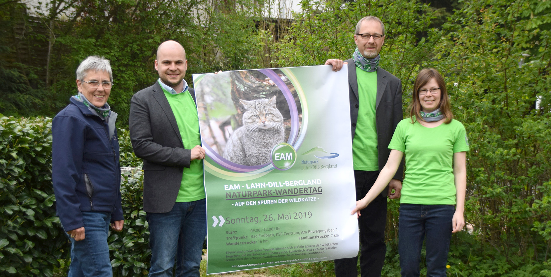 Vier Personen mit grünen T-Shirts halten ein Plakat für den Naturpark-Wandertag im Freien, umgeben von Pflanzen.