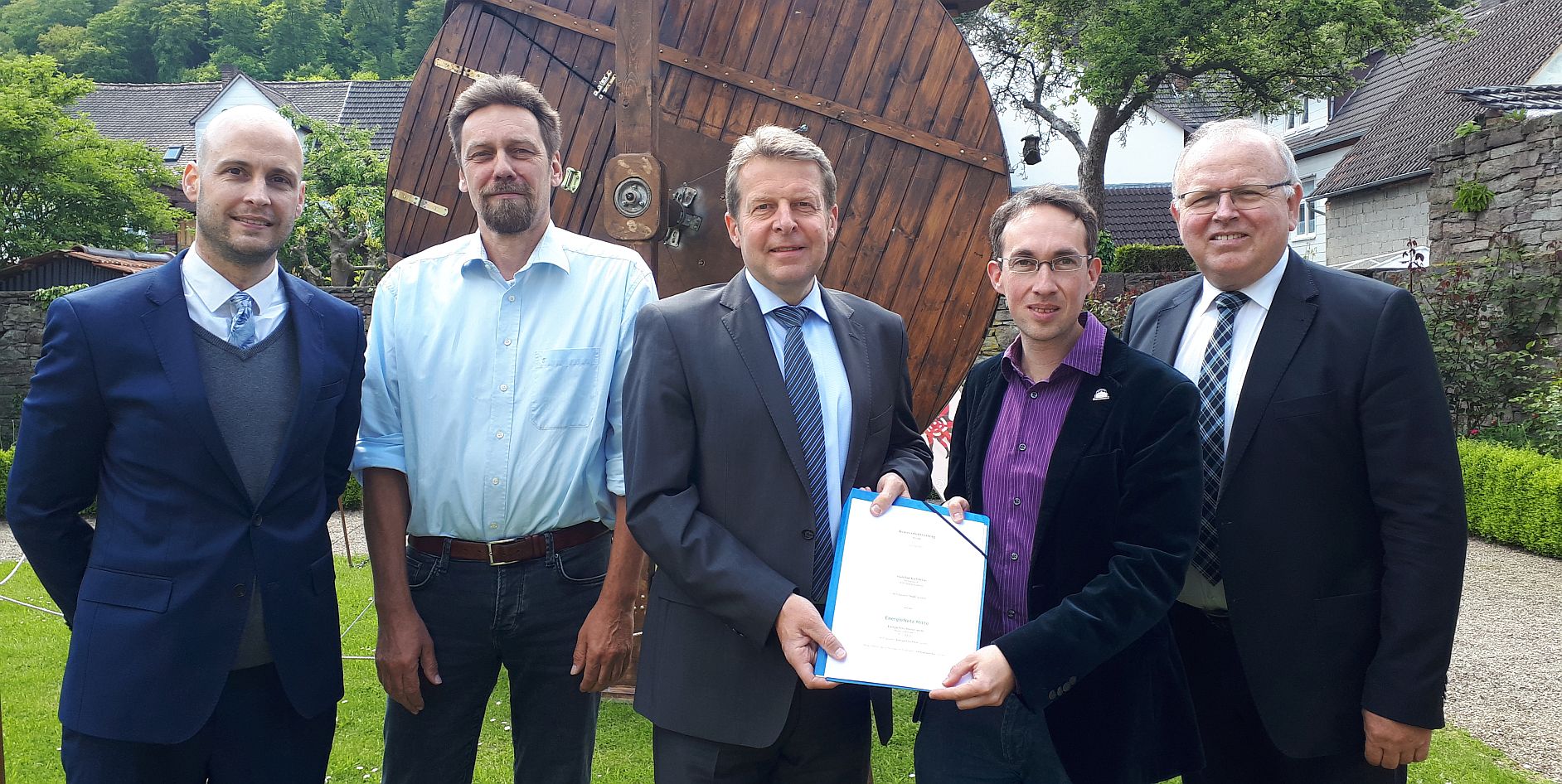 Fünf Männer stehen in einem Garten, lächeln und halten ein Dokument in der Hand. Im Hintergrund eine große Holzstruktur.