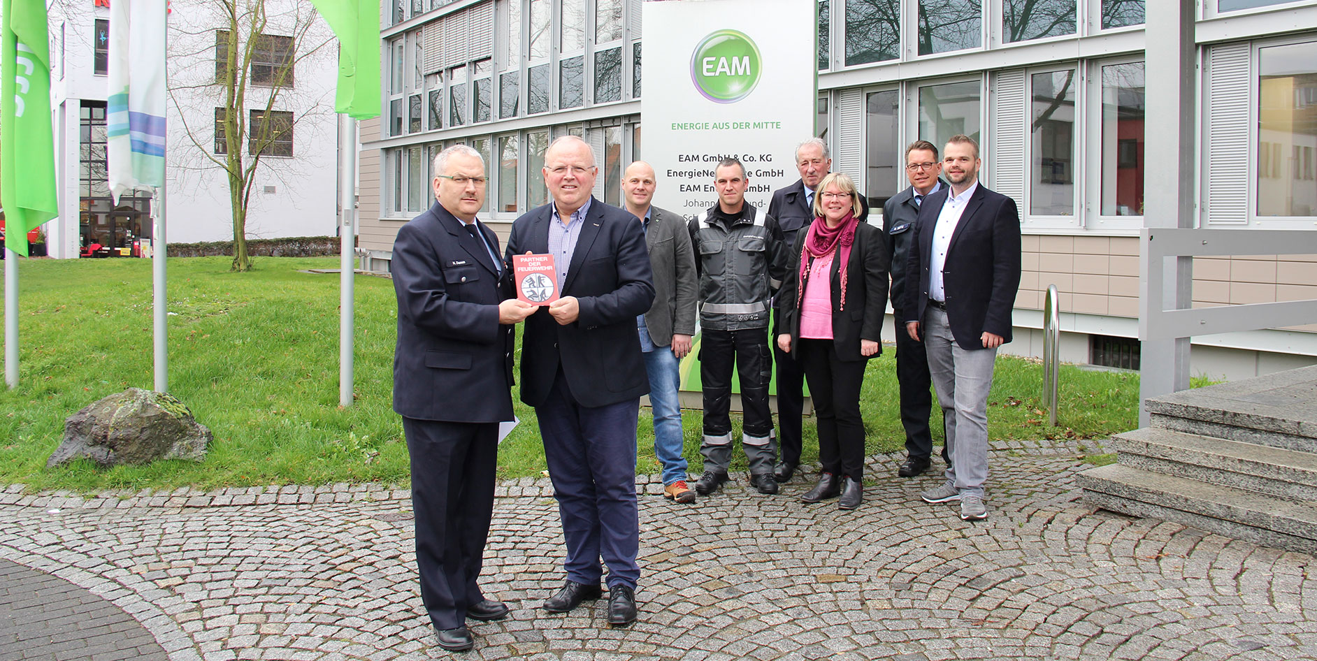 Eine Gruppe von Personen steht vor einem Gebäude mit EAM-Schild und hält ein Buch. Führende Positionen sind zu sehen.