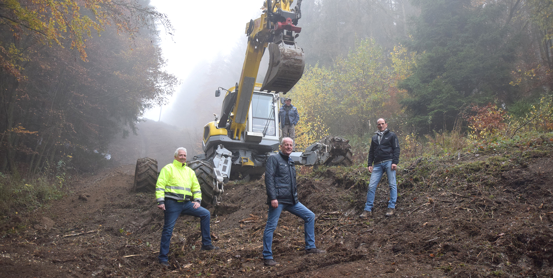 Vier Männer stehen vor einem Bagger in einem nebligen Waldgebiet mit herbstlichen Bäumen im Hintergrund.