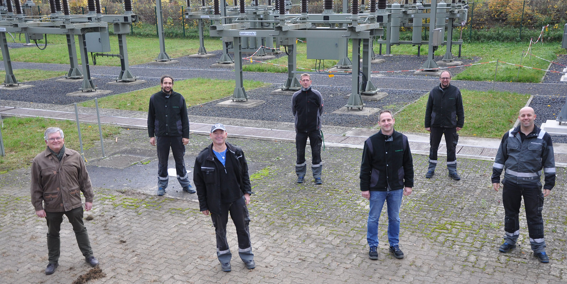 Gruppenfoto von sieben Mitarbeitenden in Arbeitskleidung, im Hintergrund technische Anlagen auf einem Außengelände.