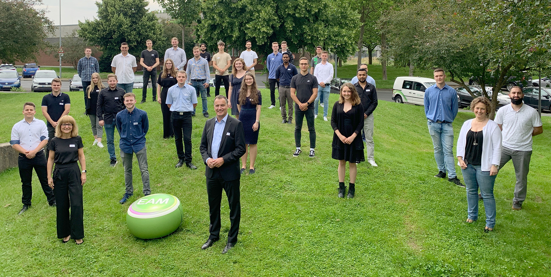 Großes Gruppenfoto der ausgelernten Auszubildenden auf einer Wiese, einige tragen Namensschilder, EAM-Logo auf einem grünen Ball im Vordergrund.