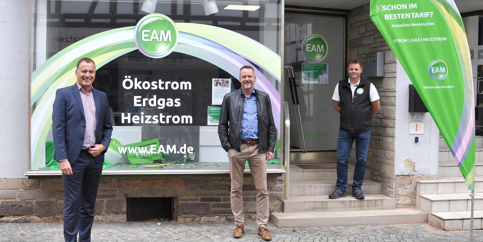 Drei Männer stehen vor dem EAM-Kundenservicebüro in Melsungen, umgeben von Werbematerialien für Ökostrom, Gas und Heizstrom.
