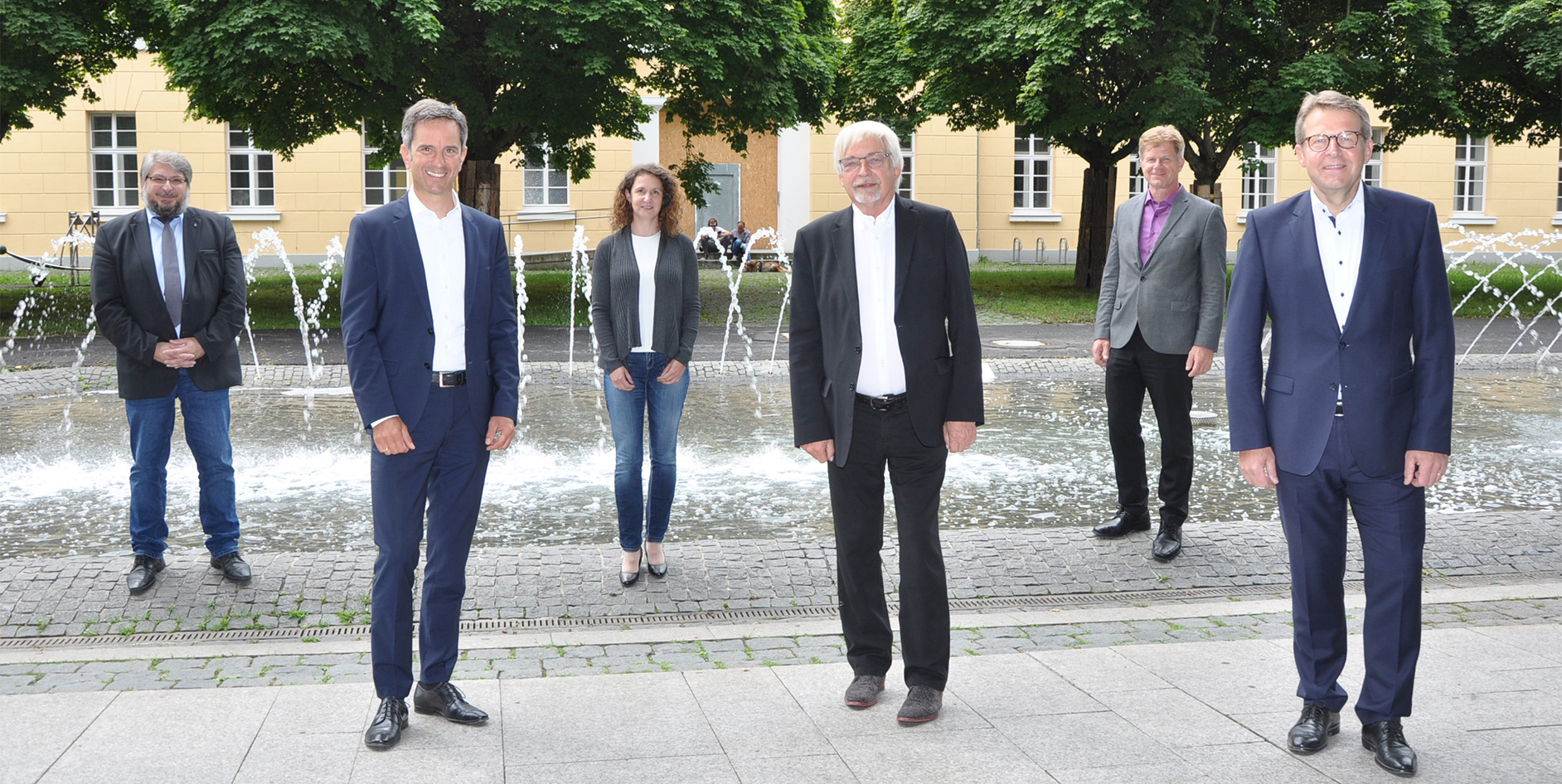 Sechs Personen stehen vor einem Brunnen in einem parkähnlichen Bereich, umgeben von Bäumen und einem Gebäude im Hintergrund.