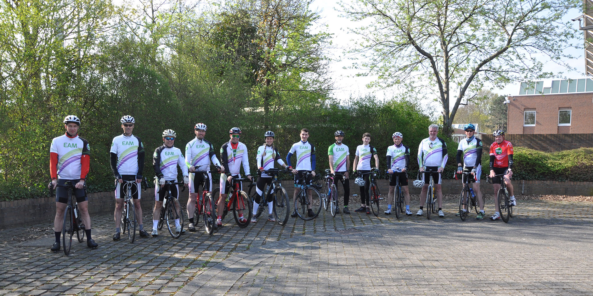 Eine Gruppe von Radfahrern in einheitlichen Trikots steht auf einem gepflasterten Platz umgeben von Bäumen.