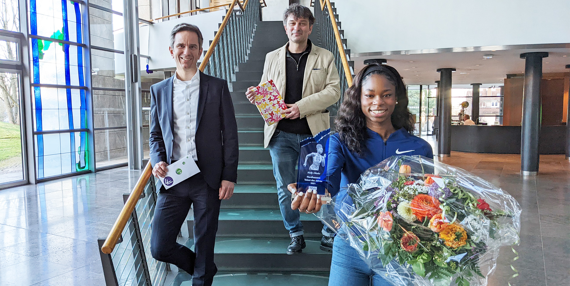 Drei Personen stehen auf einer Treppe in einem modernen Gebäude. Eine Person hält eine große Blumenausgabe und einen Preis.