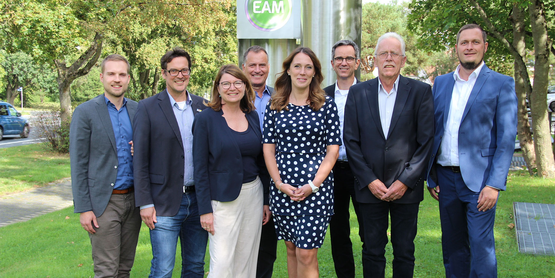 Gruppenbild vor einem EAM-Schild, umgeben von Bäumen und Rasen. Alle lächeln und stehen auf einem grünen Grundstück.