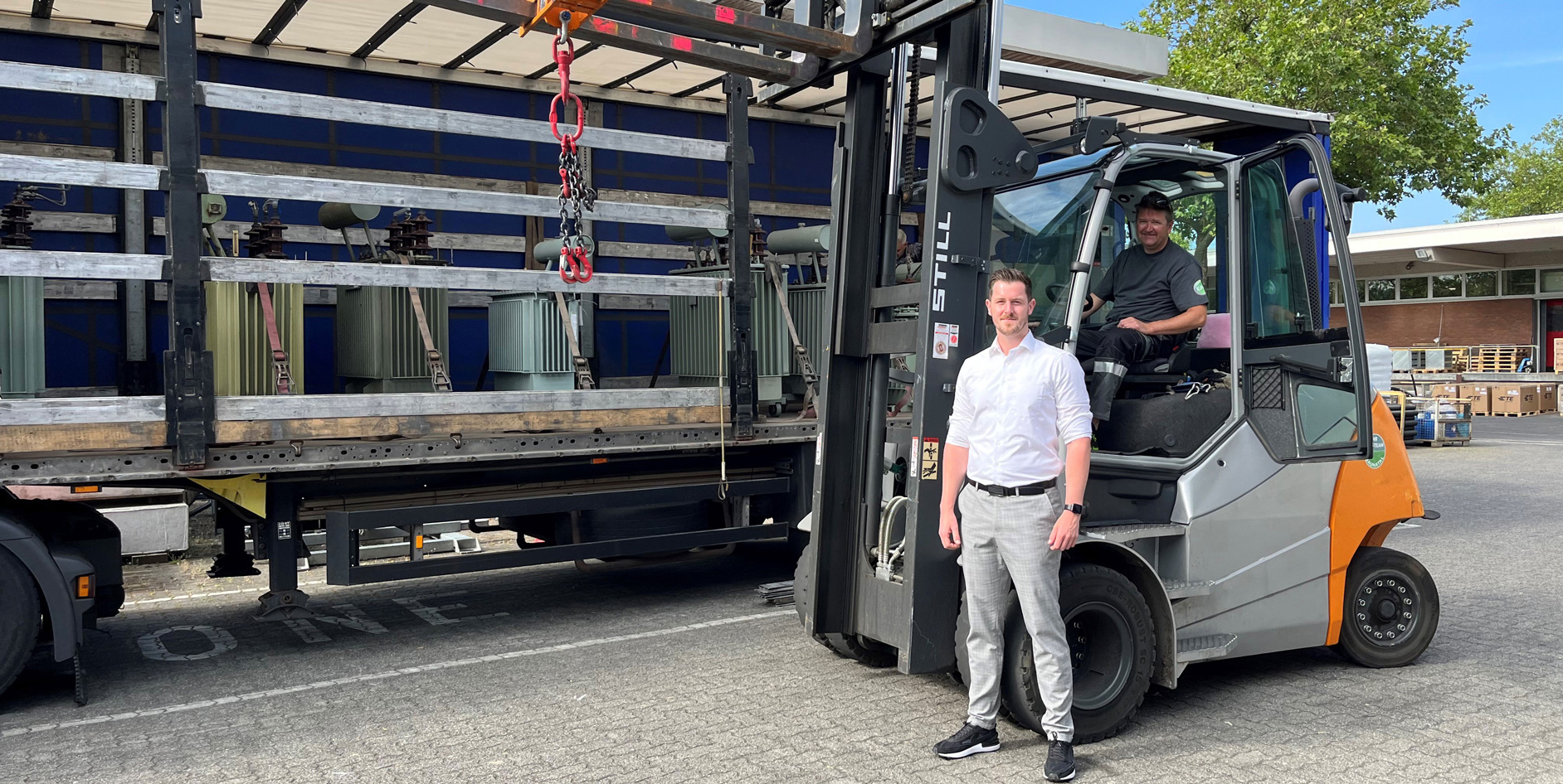 Ein Mann steht neben einem Gabelstapler, während ein Fahrer im Fahrzeug sitzt, im Hintergrund sind Paletten und einen Transporter sichtbar.