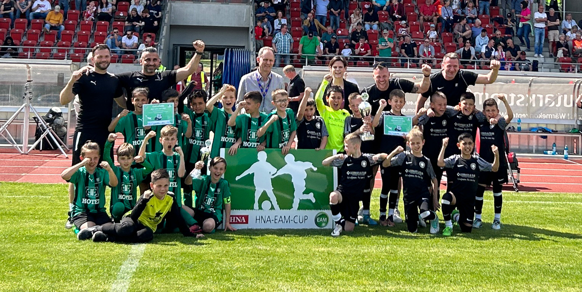 Gruppensfoto von jungen Fußballspielern mit Pokalen, mit jubelnden Coaches und einer Trophäe beim HNA-EAM-Cup.