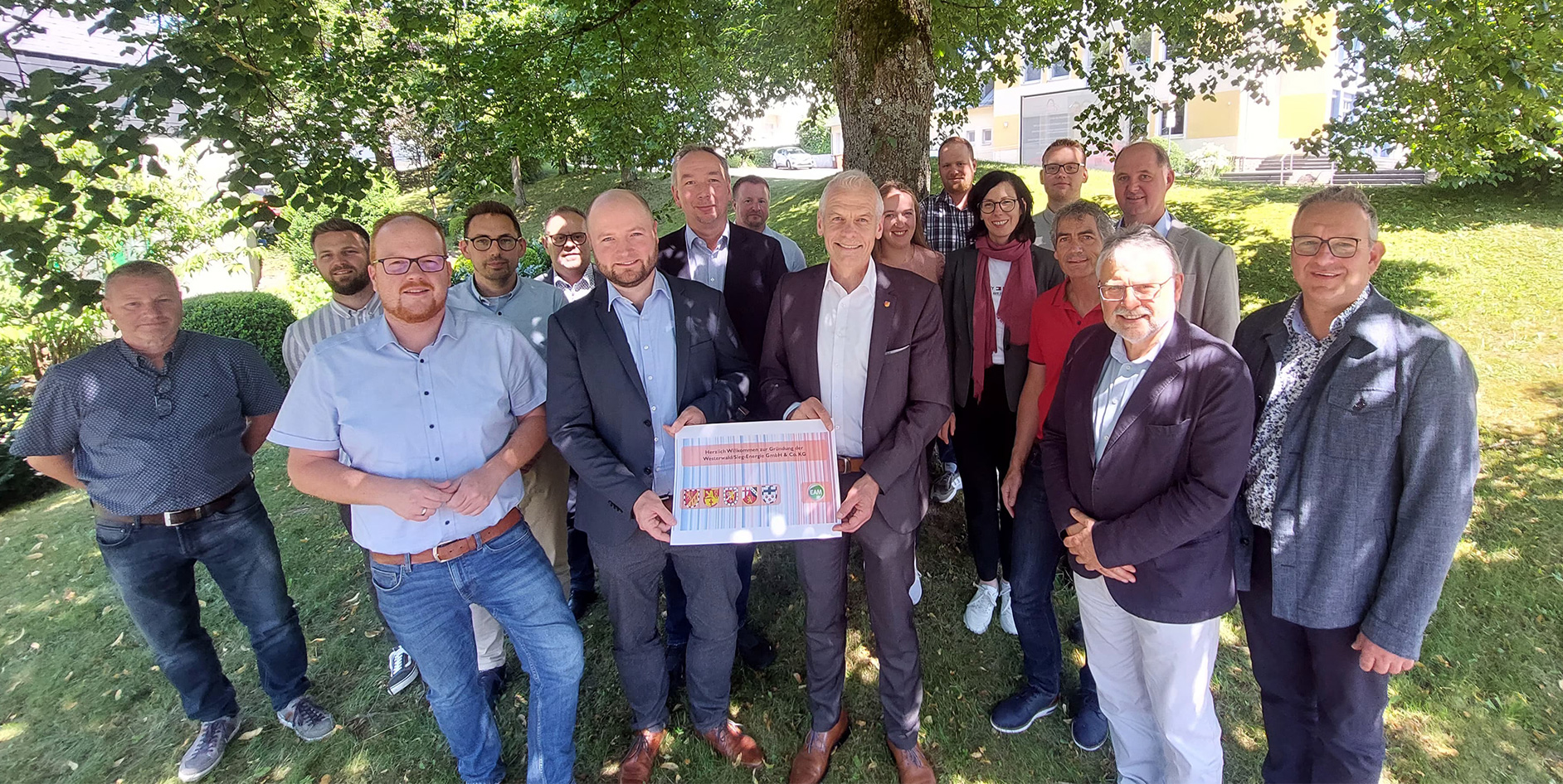 Gruppenfoto von Personengruppe im Freien, einige halten ein Schild mit den Farben und Wappen von Unternehmen und Organisationen.