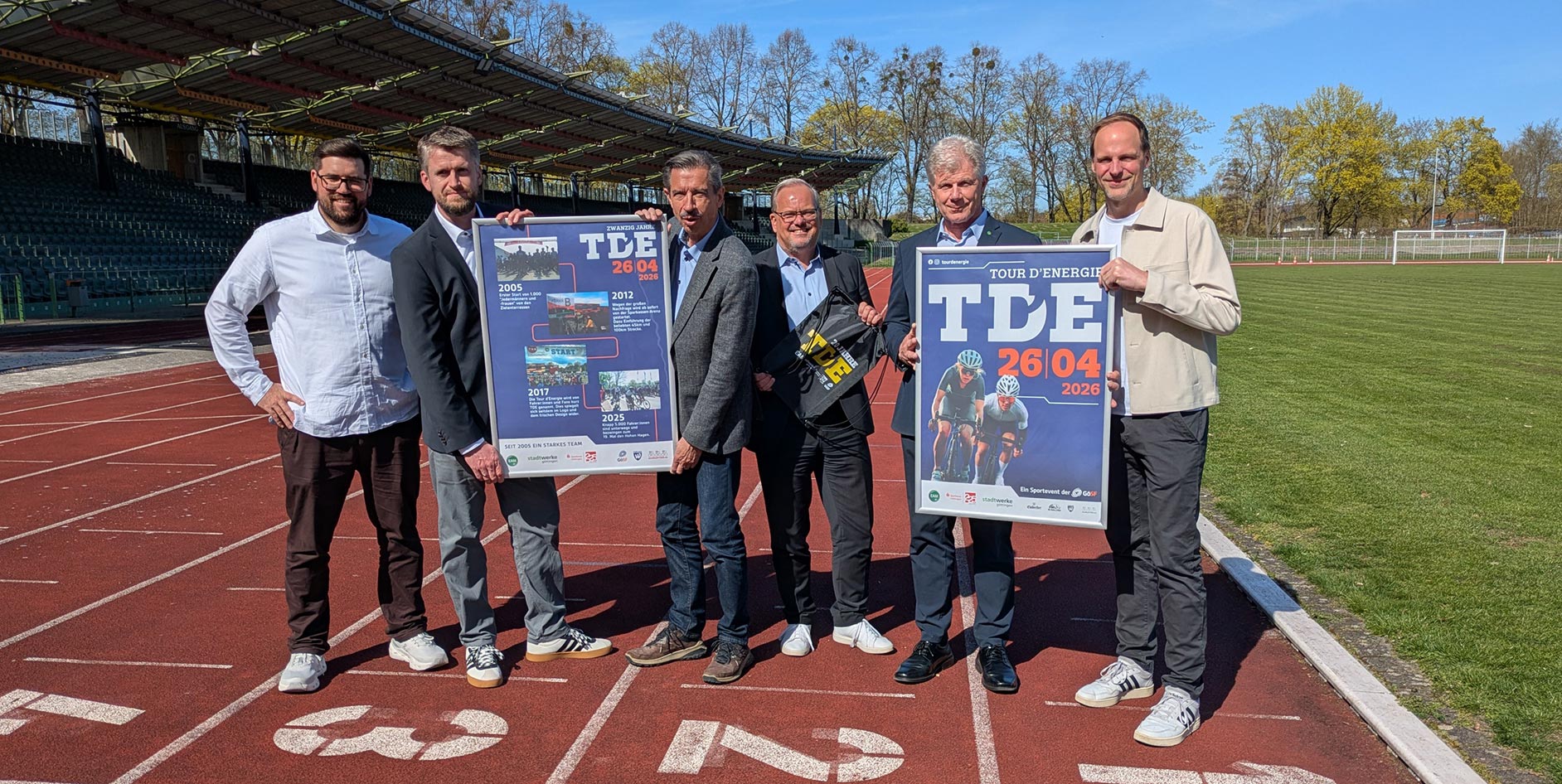 Sechs Personen stehen auf einer Laufbahn in einem Stadion und halten Veranstaltungsplakate in die Kamera, darunter ein Plakat zur „Tour d’Energie“ mit dem Datum 26.04.2026; im Hintergrund sind Tribüne, Rasenfläche und Bäume bei sonnigem Wetter zu sehen.
