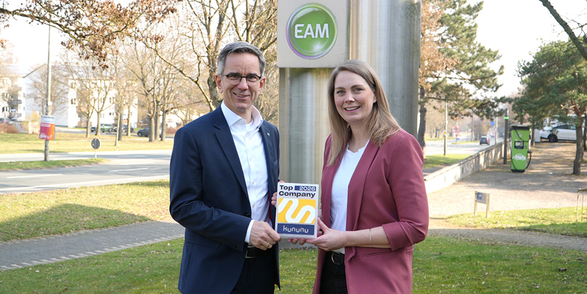 Zwei Personen stehen im Freien vor einem EAM‑Schild und halten gemeinsam eine Auszeichnung mit der Aufschrift „Top 2026 Company – kununu“. Im Hintergrund sind eine Straße, Bäume, ein Gehweg und eine Ladeinfrastruktur für Elektrofahrzeuge zu sehen.