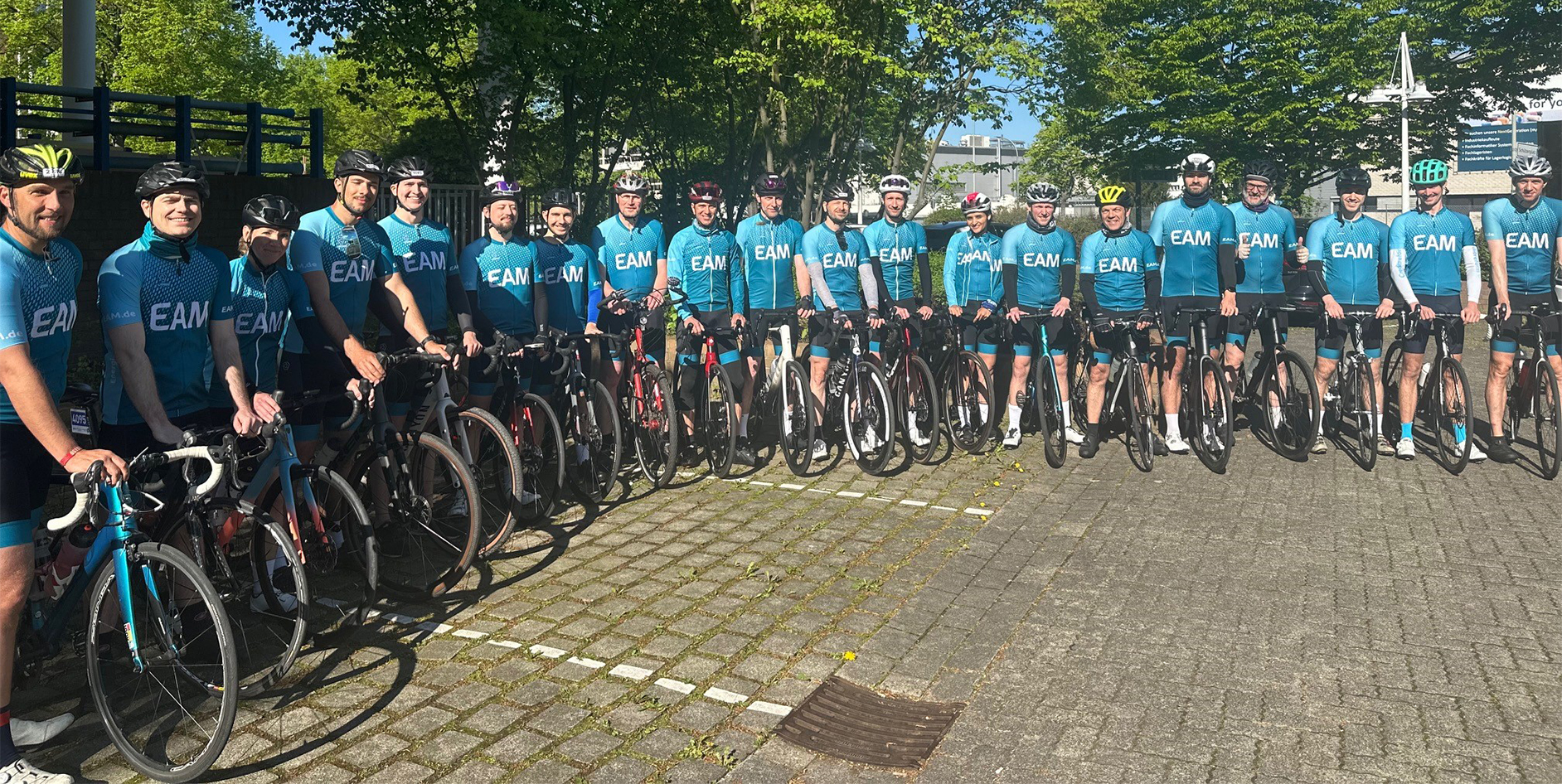 Eine Gruppe Radfahrer in blauen EAM-Trikots und Helmen stehen mit ihren Rennrädern auf einem gepflasterten Platz.