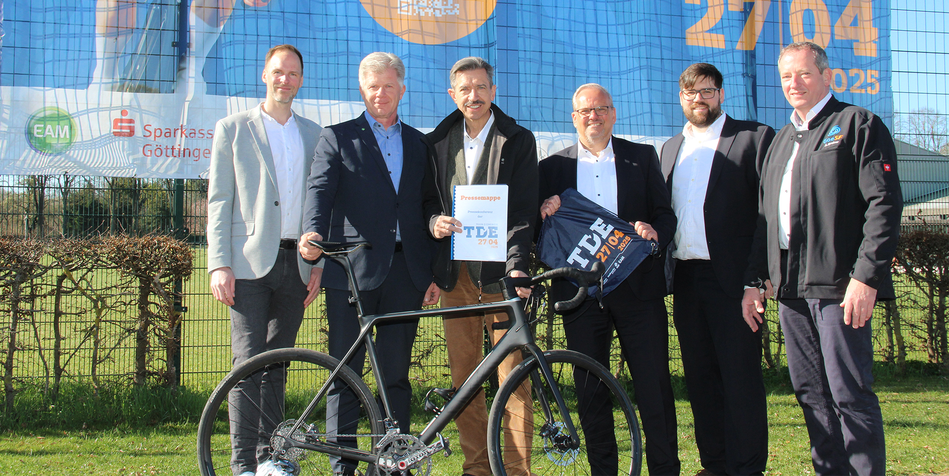 Sechs Personen stehen vor einem großen Banner, vor ihnen steht ein modernes Rennrad und sie halten eine TDE-Urkunde.
