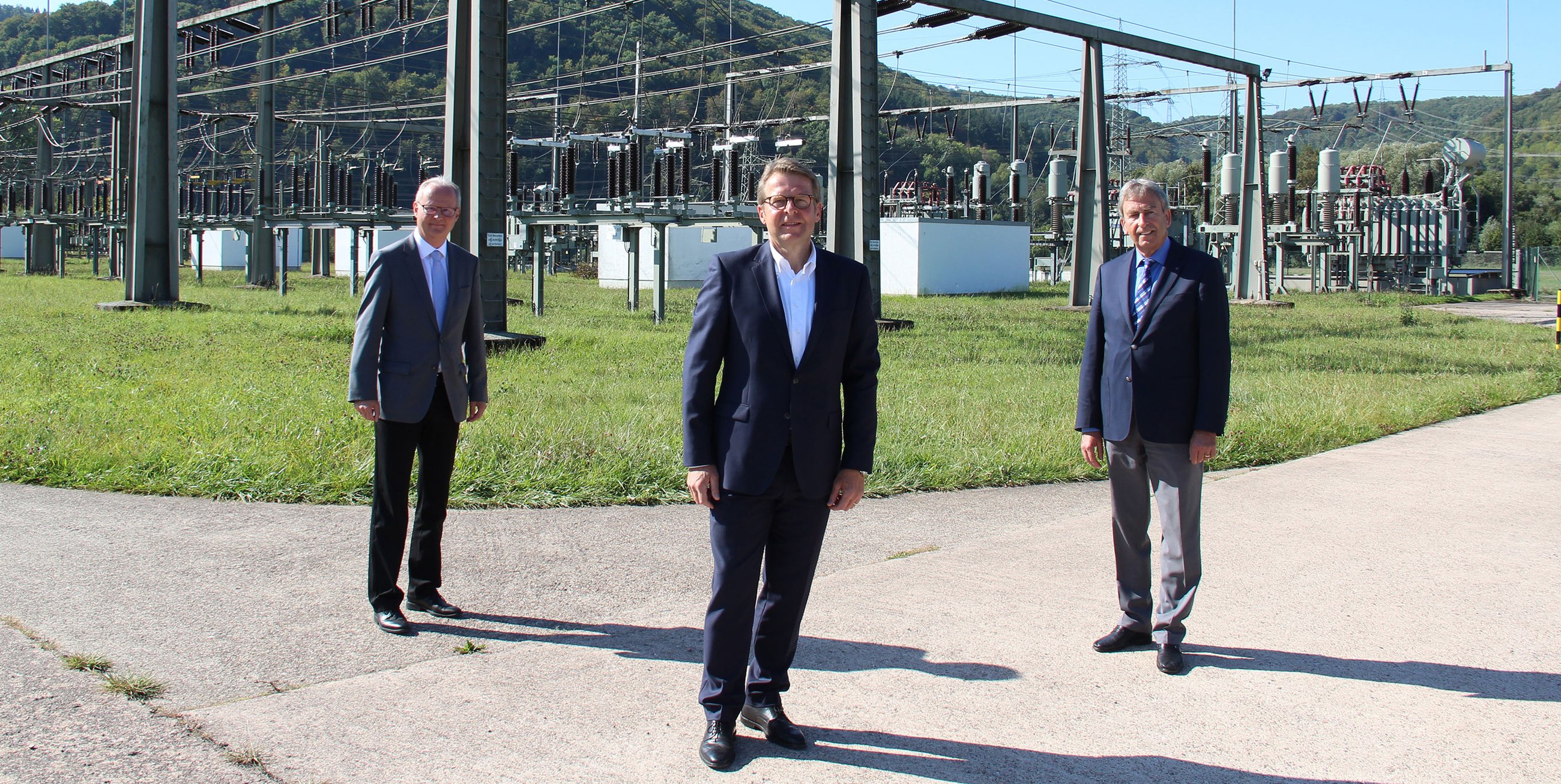 Drei Männer in formeller Kleidung stehen vor einer elektrischen Umspannstation in einem grünen Landschaftsbereich.
