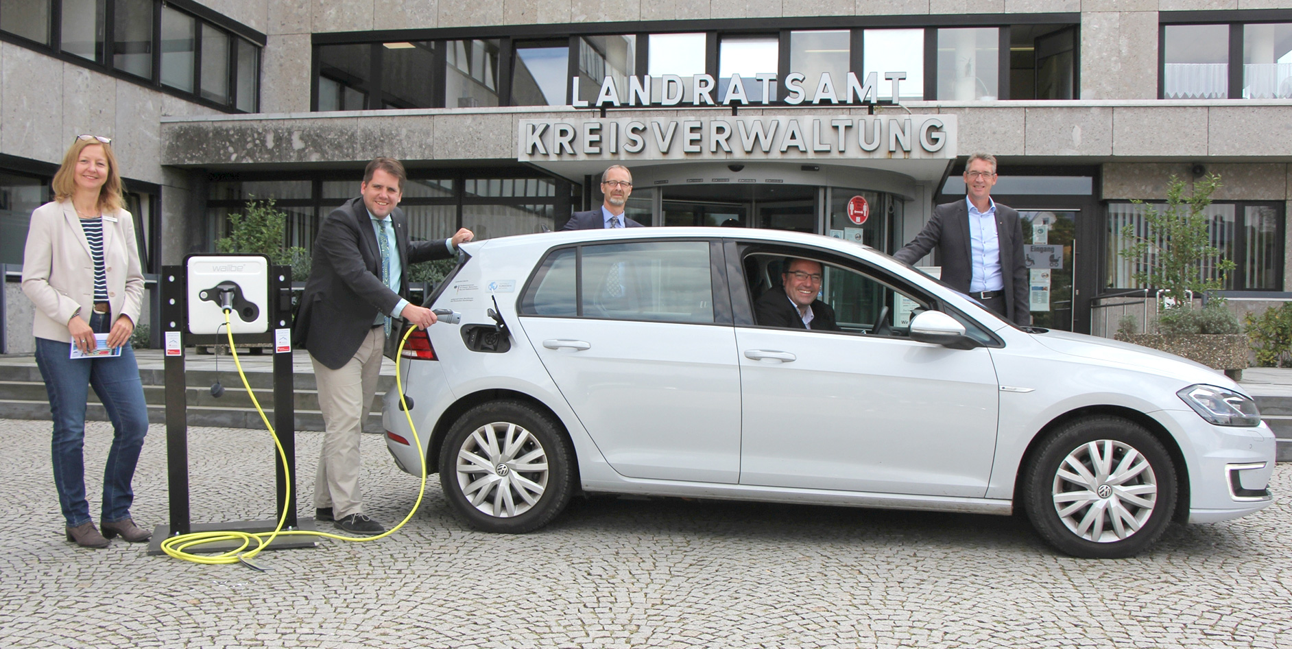 Der Landkreis Marburg-Biedenkopf fördert E-Ladestationen Fünf Personen stehen an einer Elektroladesäule neben einem Elektroauto vor einem Verwaltungsgebäude.