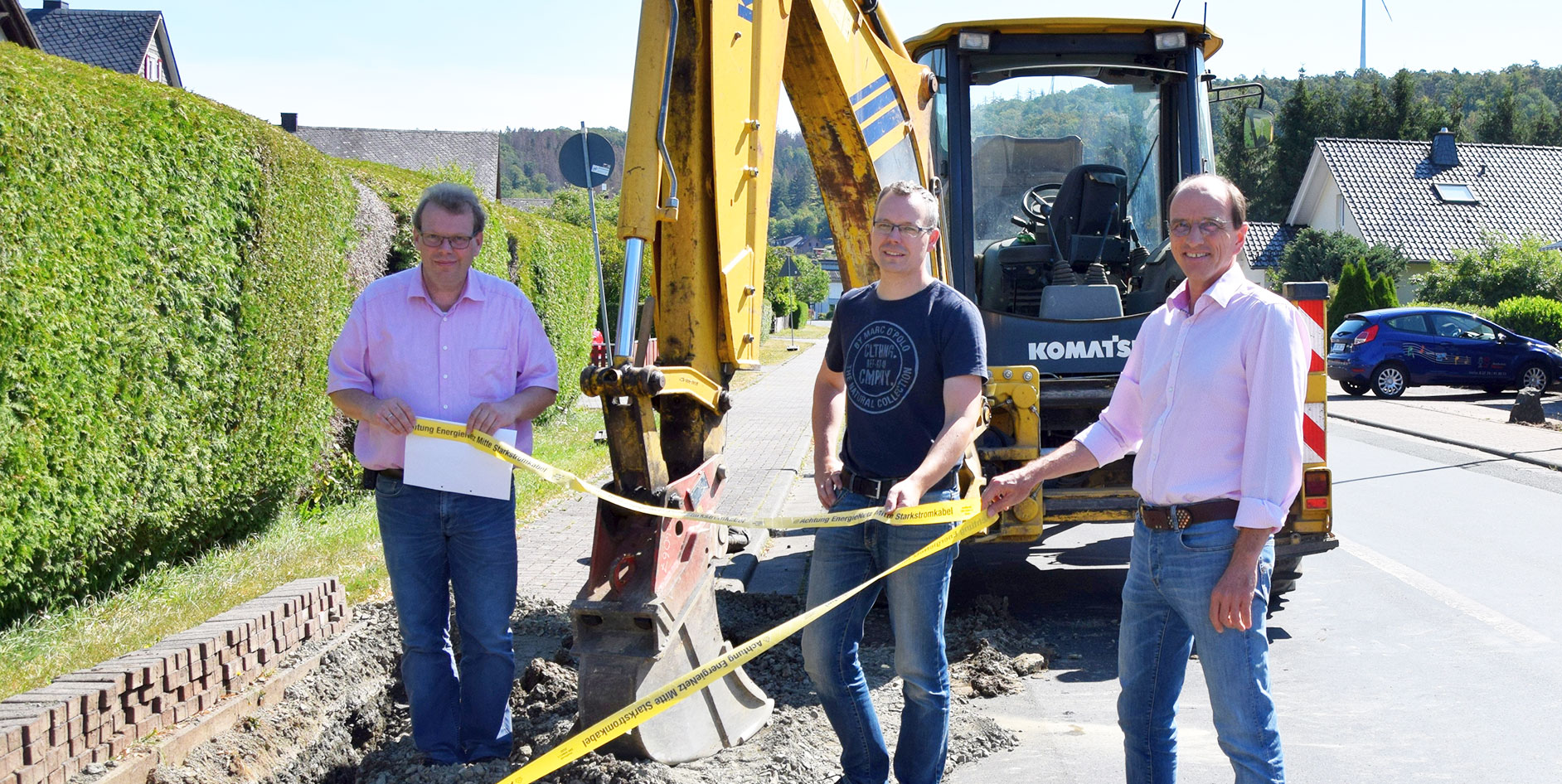Drei Männer stehen an einer Baustelle mit einem Bagger und halten ein Sicherheitsband.