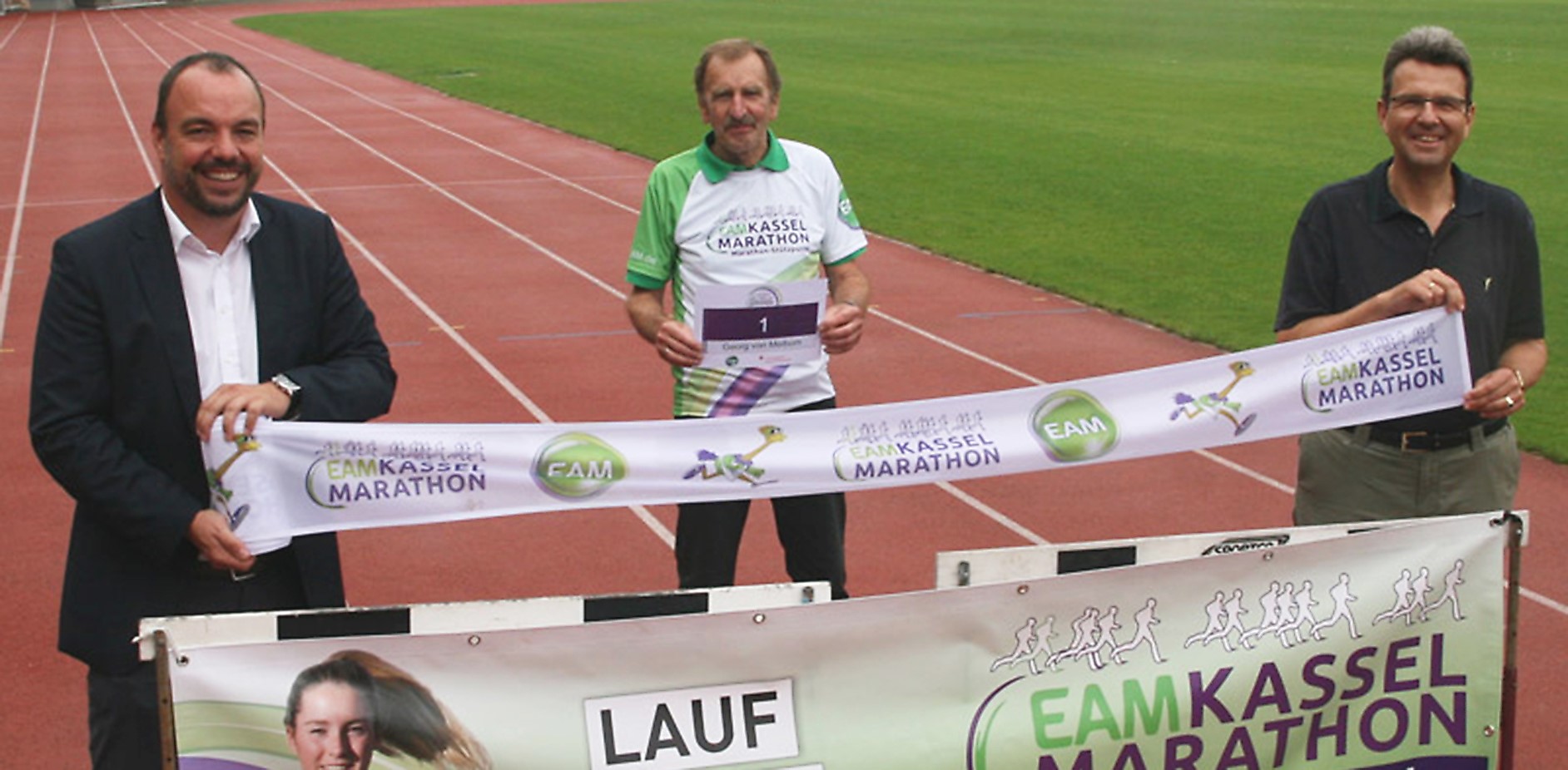 Drei Personen stehen auf einer Laufbahn, halten ein Banner des Kassel Marathons und präsentieren Medaillen.