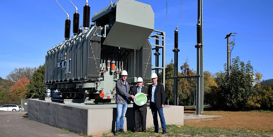Neuer Transformator für das Umspannwerk Ottrau Drei Personen in Arbeitskleidung mit einem Energiemanagement-Schild vor einem Transformator im Freien.