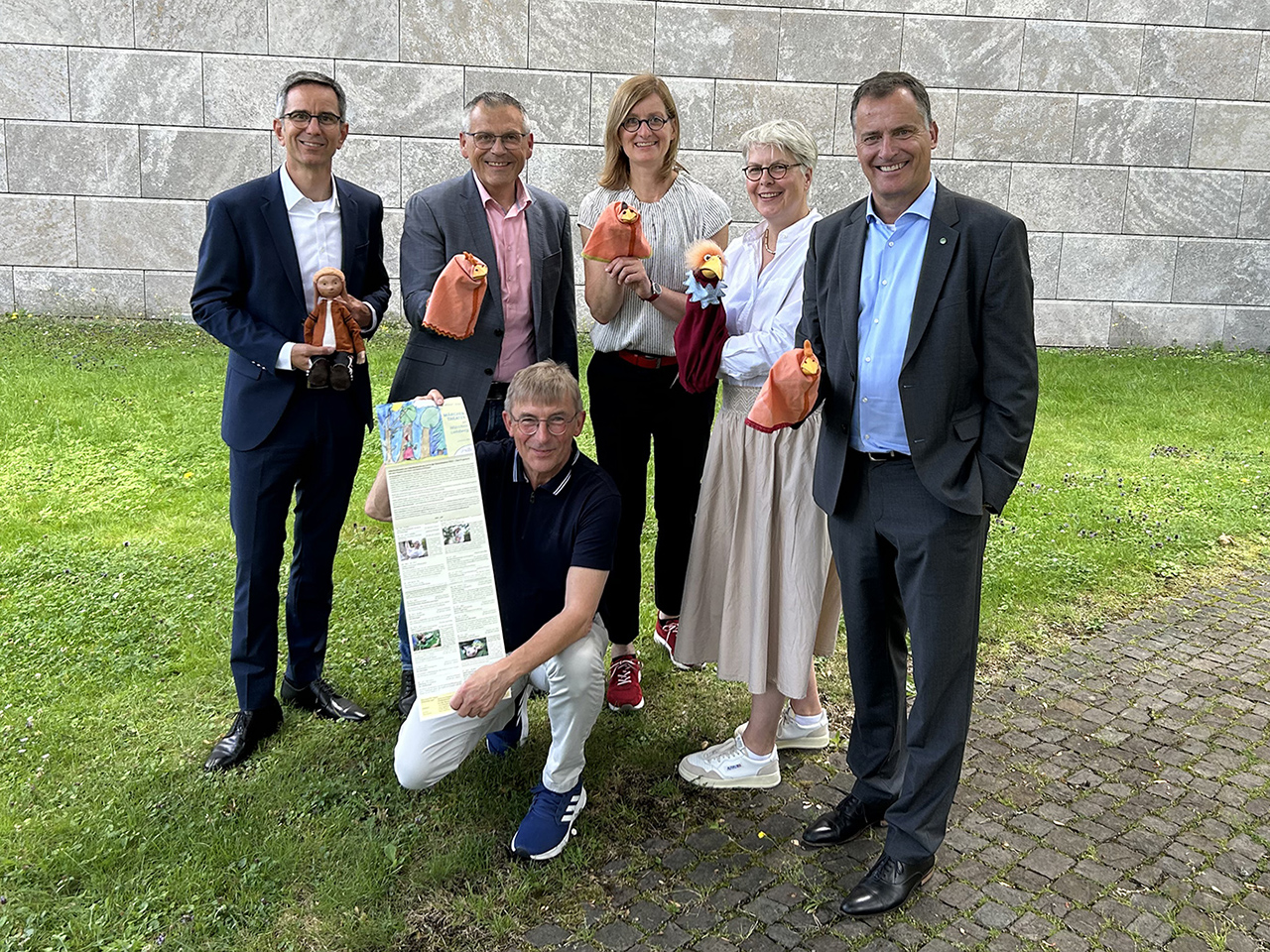 Sechs Personen stehen auf einer Wiese, halten Stoffpuppen und ein Plakat, mit einer Steinmauer im Hintergrund.
