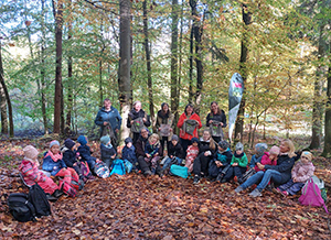 Gruppe von Kindern und Erwachsenen im Herbstwald, umgeben von buntem Laub, sitzend auf dem Boden.