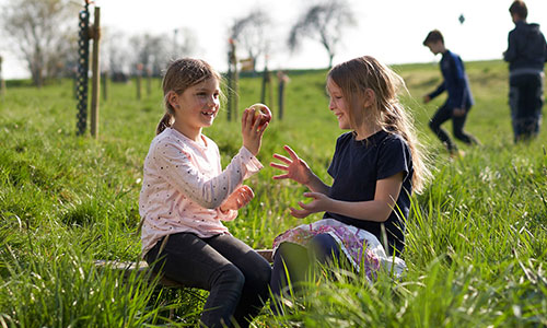 Zwei Mädchen sitzen im Gras und spielen mit einem Apfel, während andere Kinder im Hintergrund spielen.