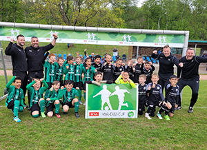 Junge Fußballmannschaft feiert ihren Erfolg beim Turnier, umgeben von Trainern und einem Banner.
