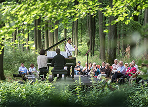 Zwei Musiker spielen an einem Klavier im Wald, während ein Publikum aus Sitzenden zuhört. Grün umgebene Natur.