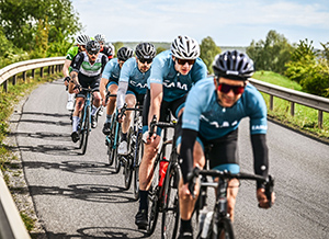 Sechs Radfahrer in blauen Trikots fahren auf einer kurvenreichen Straße, umgeben von grüner Landschaft.