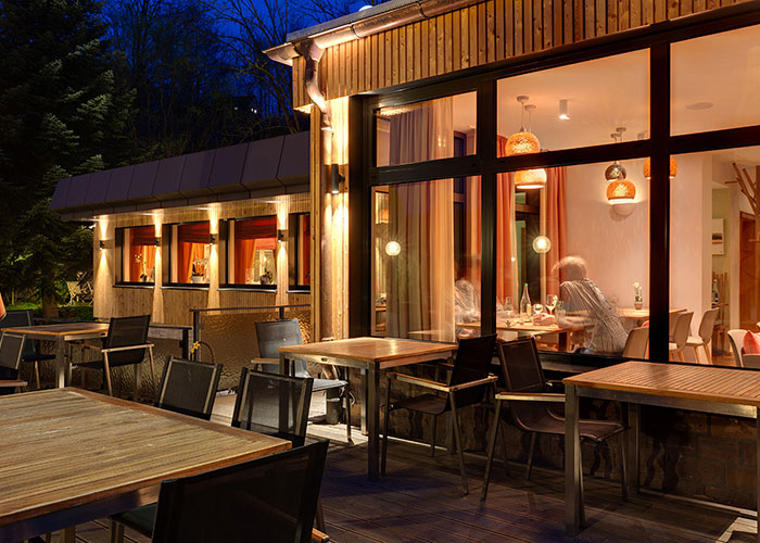 Beleuchtete Außenterrasse eines Restaurants bei Nacht, mit Holzwänden und Tischen. Gäste sitzen drinnen sichtbar.
