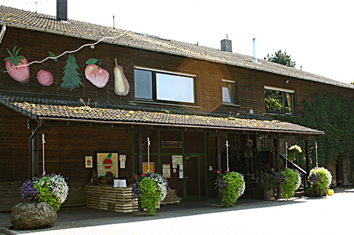Holzgebäude mit bunten Gemälden von Obst und Gemüse, umgeben von blühenden Pflanzen und einem einladenden Eingangsbereich.