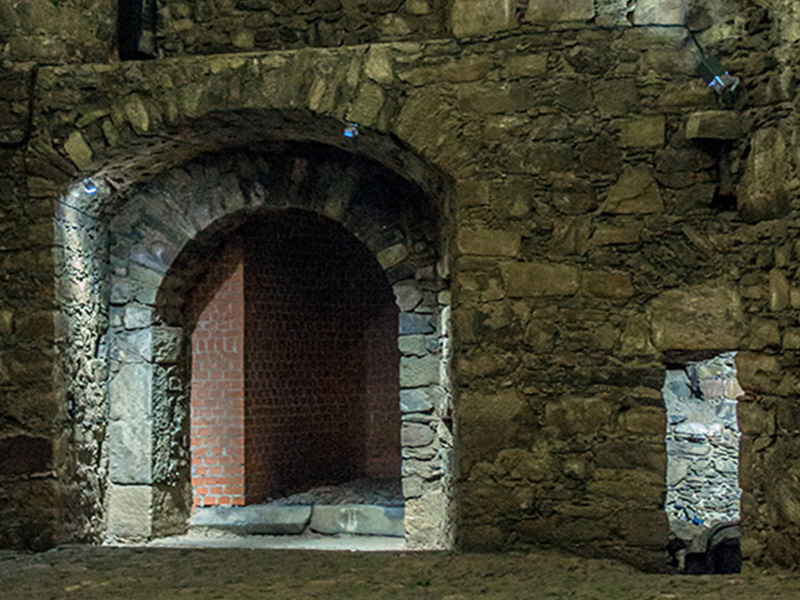 Alter Steinbogen mit zwei Öffnungen in einer historischen Mauer, beleuchtet von oben, dunkler Hintergrund.