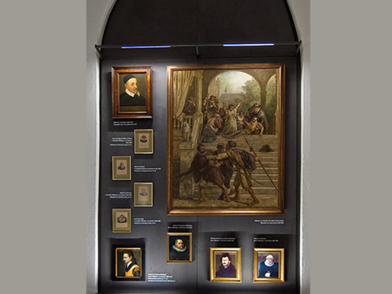 Wand mit mehreren gerahmten Porträts und einem großen Gemälde, das eine historische Szene mit Menschen darstellt.
