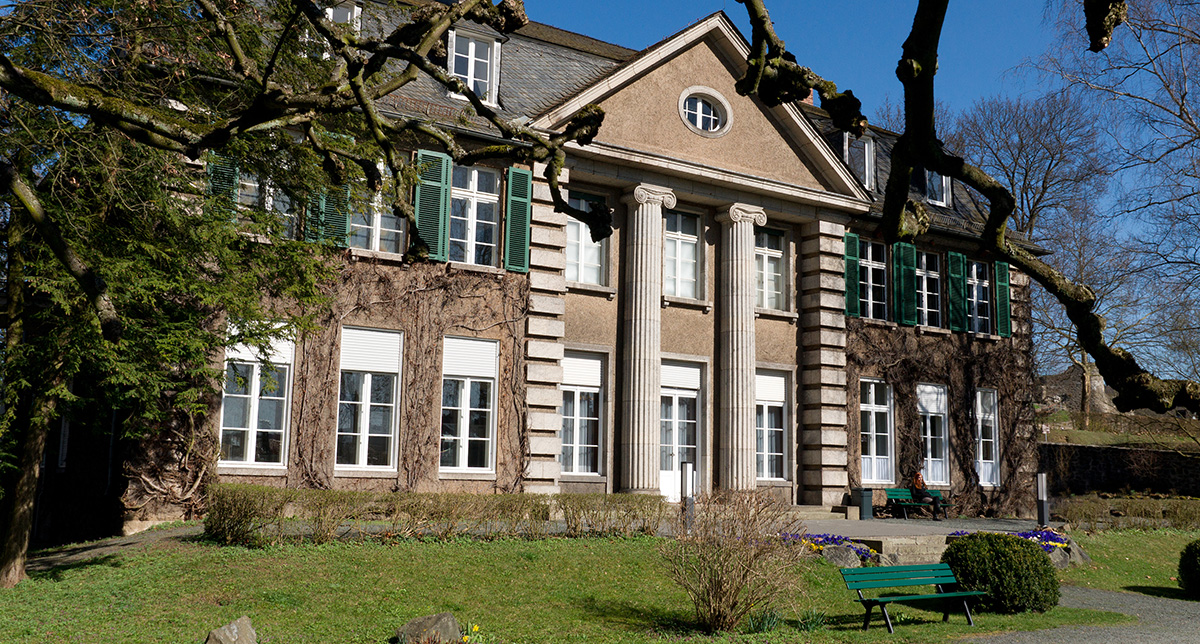 Historisches Gebäude mit grünen Fensterläden, Säulen und umgebender Vegetation, unter blauem Himmel.