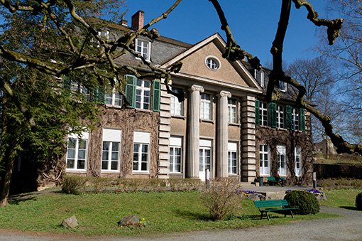 Historisches Gebäude mit Säulen, umgeben von Bäumen und einer Wiese, im Hintergrund blauer Himmel.