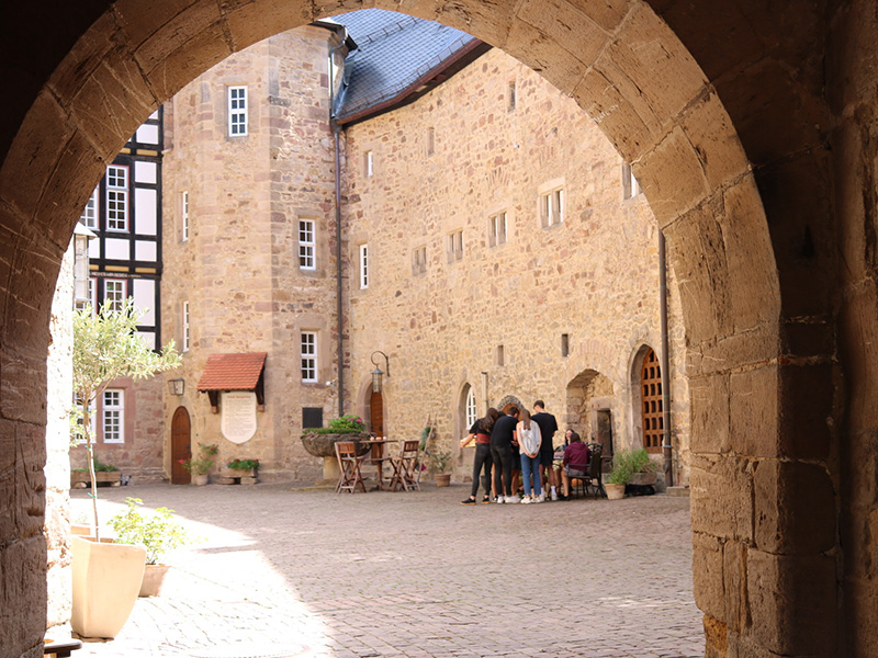Innenhof eines historischen Gebäudes mit Personen, die an einem Tisch stehen, umgeben von Steinen und Pflanzen.