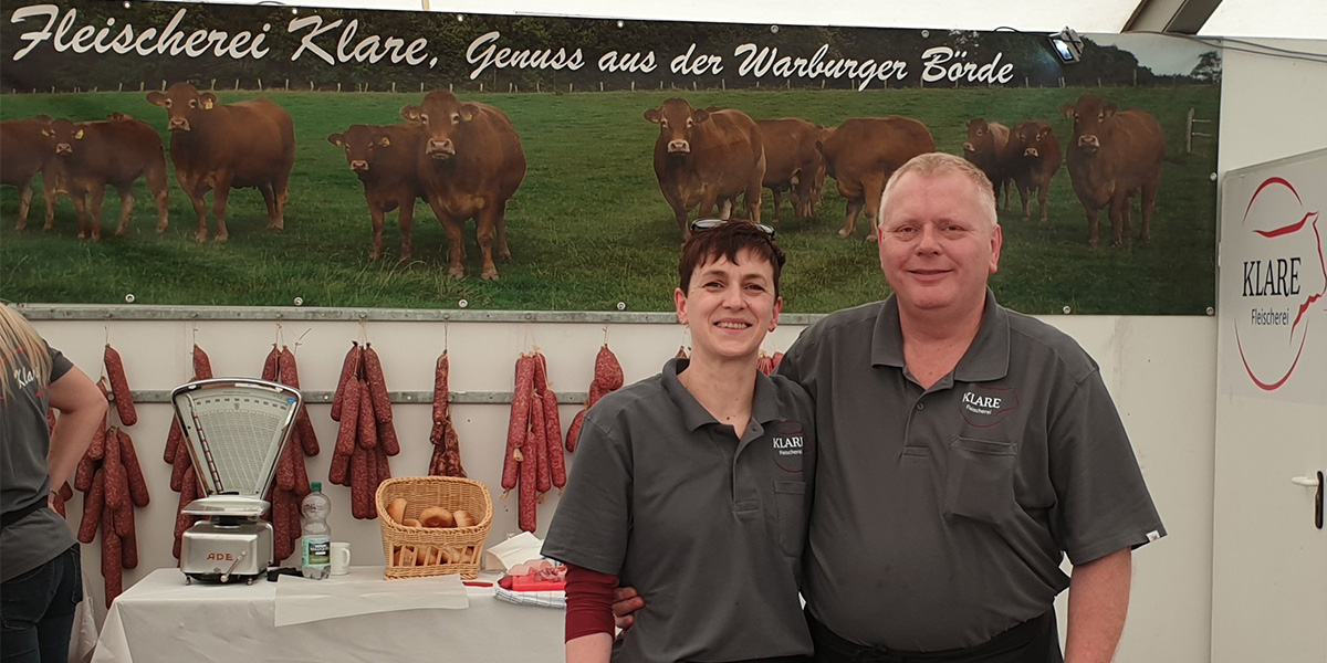 Zwei Personen lächeln in die Kamera und stehen vor einer Theke an der Fleisch verkauft und abgewogen wird.