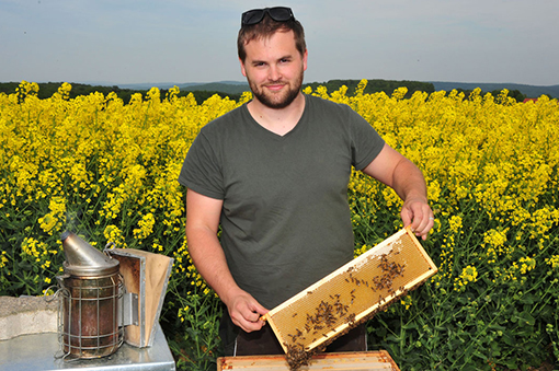 Mann mit Bienenstock vor einem Rapsfeld, hält eine Bienenwabe in der Hand und trägt Schutzbrille.
