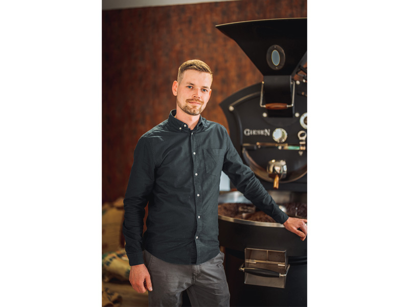 Mann in dunkler Hemd steht neben einer Kaffeeröstmaschine in einem modernen Café.