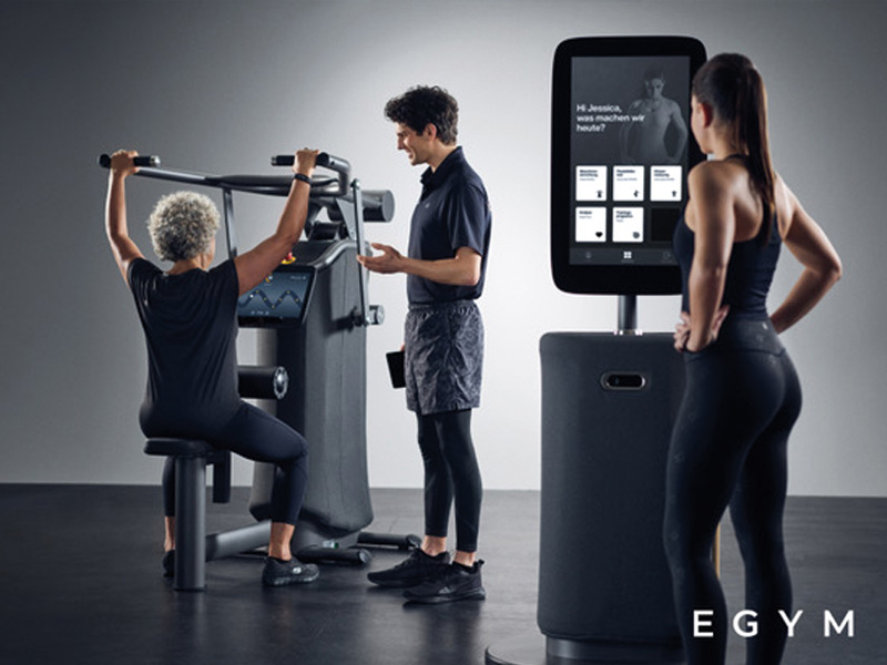 Drei Personen im Fitnessstudio: eine trainiert an einem Gerät, ein Trainer gibt Anweisungen, ein weiteres Gerät im Hintergrund.