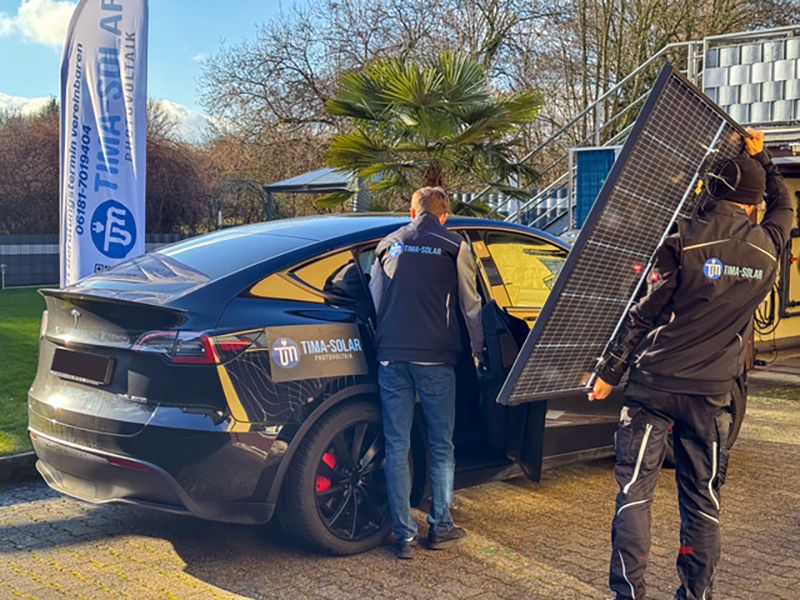 Zwei Personen am schwarzen Elektroauto von Tima Solar, eine Person trägt ein Solarpanel. Im Hintergrund eine Palmenschaltung.