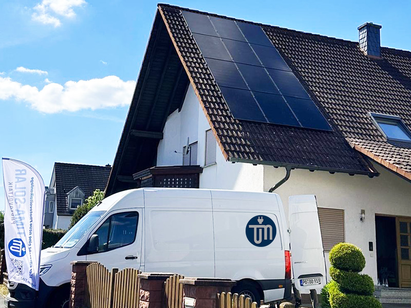 Weißer Lieferwagen vor einem modernen Haus mit Solarzellen auf dem Dach und einem Fähnchen im Vordergrund.