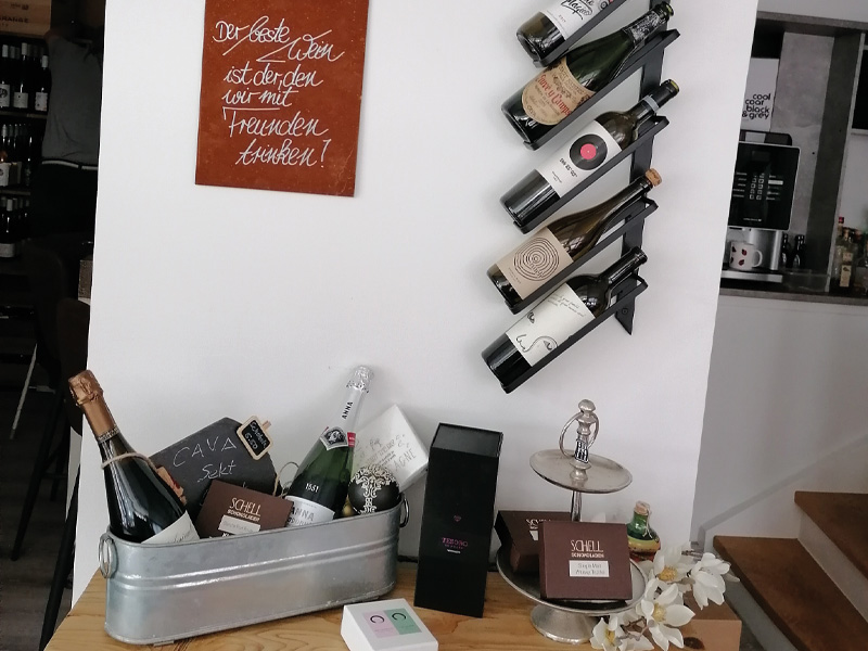 Eine Sammlung von Weinen und Sektflaschen in einem Bistro, daneben ein Holztisch mit Dekoration und einem Spruch auf der Wand.