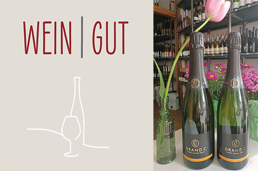 Zwei Flaschen Sekt mit dem Schriftzug "WEIN | GUT"" und einer Blume in einer Vase im Hintergrund."